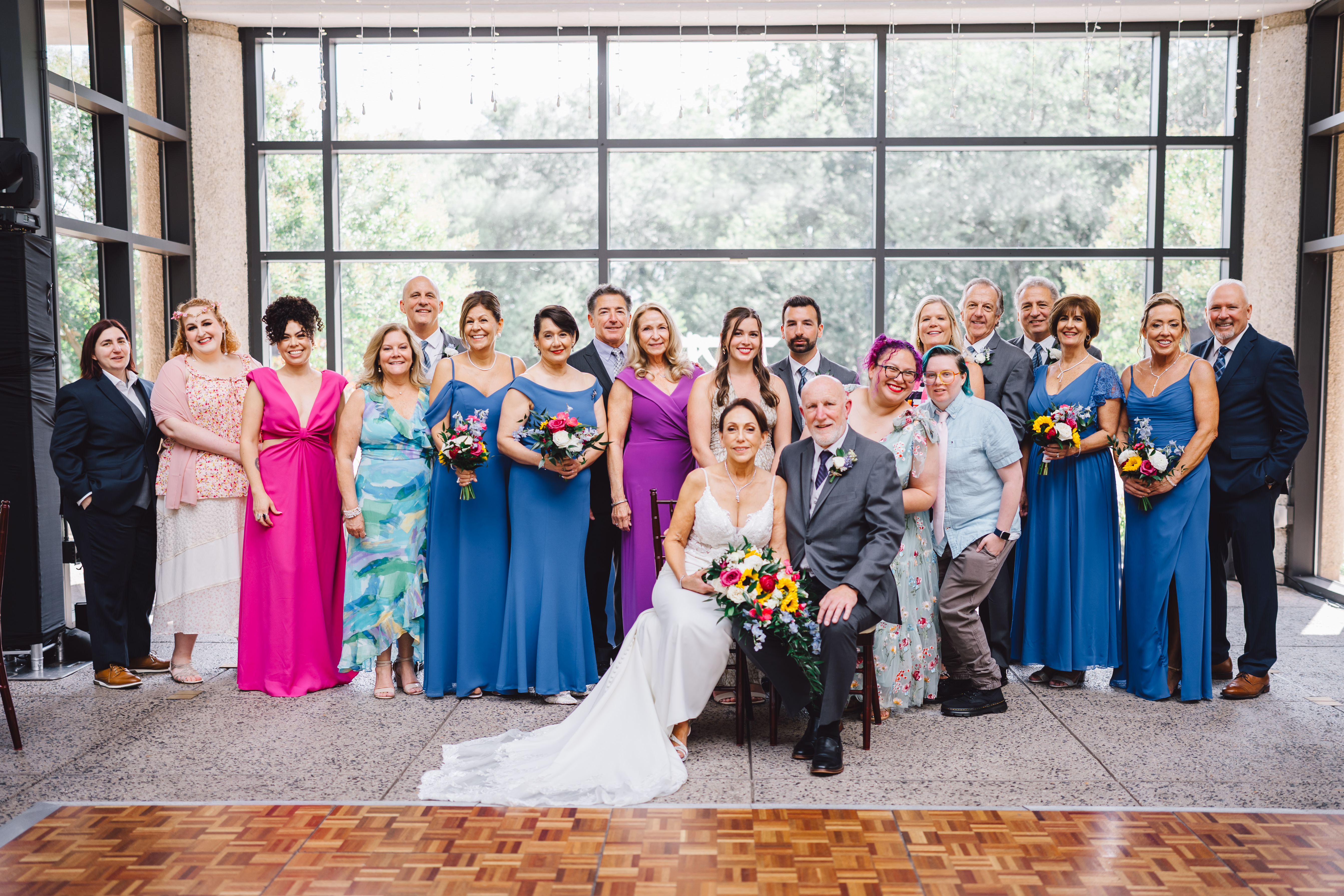 couple sitting in front of wedding party all wearing colorful clothing while in front of the Atrium's large window