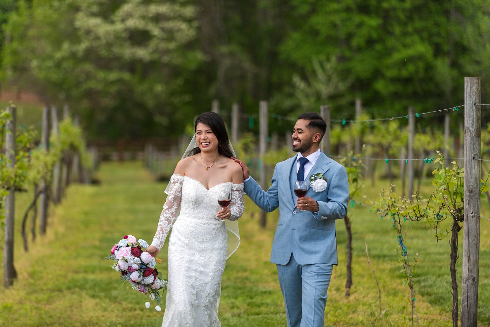 couple laughing in a vineyard holding glasses of red wine and looking into distance 