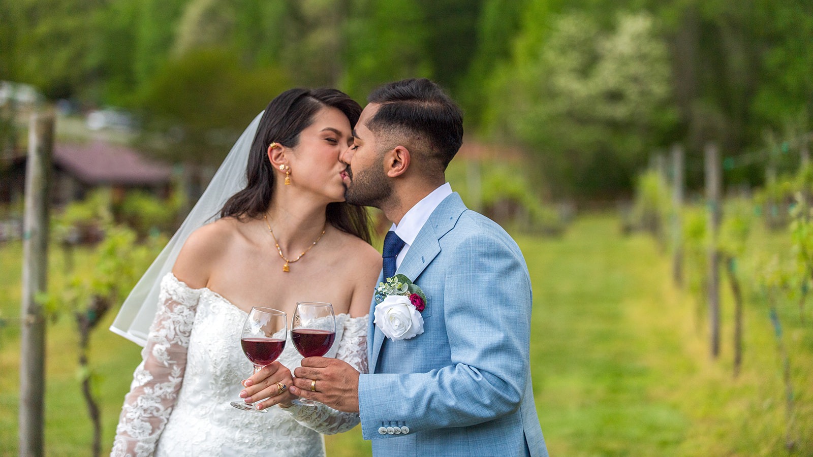 couple in the vineyard kissing while holding two glasses of red wine