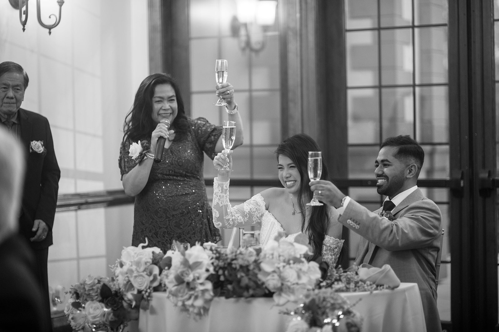 black and white photo of bride, groom, and mother raising glasses at the sweetheart table during a toast