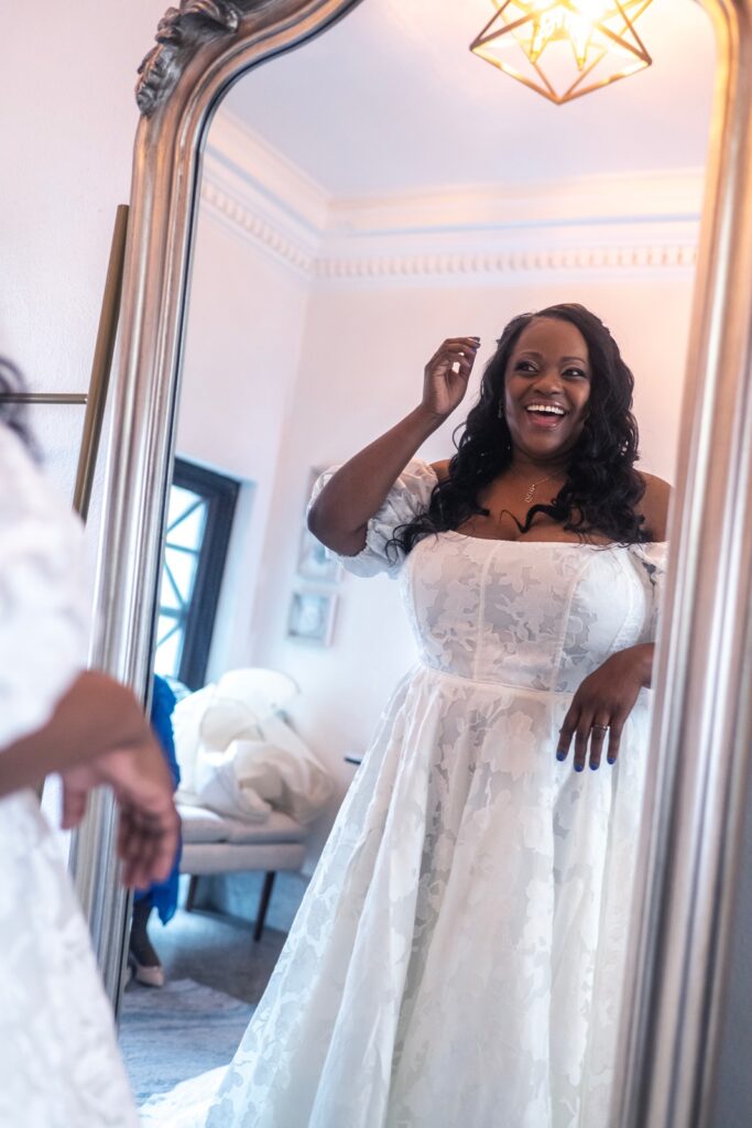 A bride in her wedding dress smiling at herself in the mirror and looking confident after her Maryland Wedding Dress Appointment.