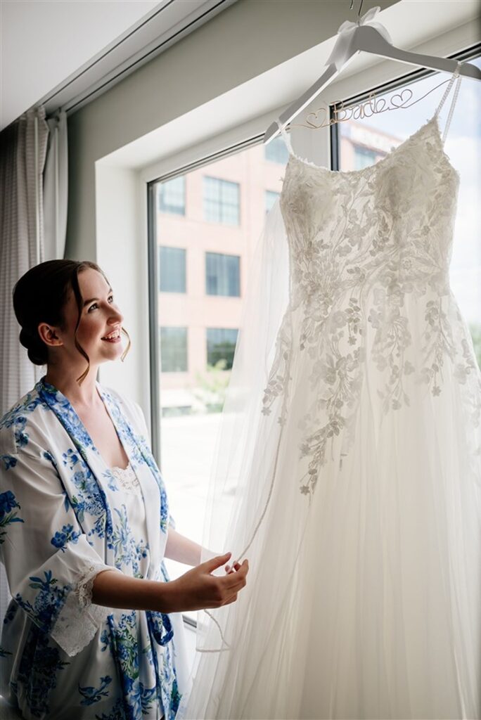 A bride admiring her wedding dress as it hangs on a hangers in front of a hanger.