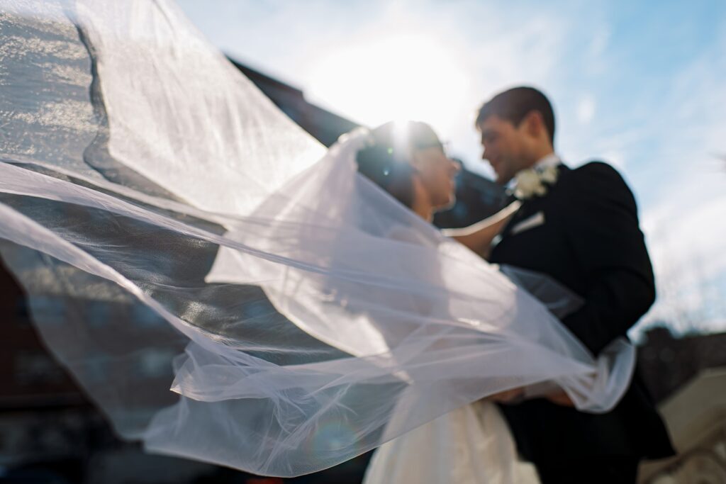 Wedding couple stands outside looking at each other while the wind blows the bride's veil.